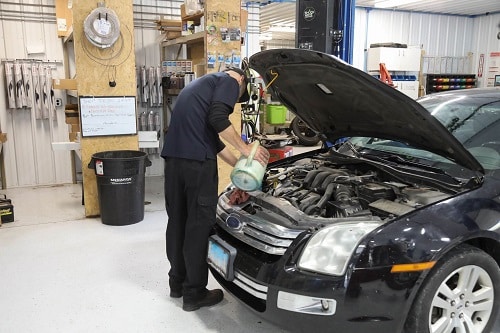 Preventative Maintenance Tips to Make a Vehicle Last Longer in Steeleville, IL | Mevert Automotive & Tire Center. Image of a male mechanic doing fluid change and refill on a black car in the auto shop garage.